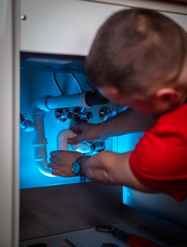 Professional plumber in a red shirt repairing under-sink drainage piping, showcasing Smart Agnes's expert plumbing and MEP services in Qatar.